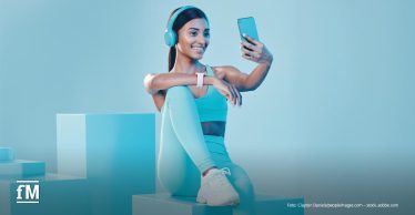 Eine Frau in heller Sportbekleidung sitzt auf einem Podest und hält ein Smartphone für ein Selfie vor sich. Sie trägt große hellblaue Kopfhörer und eine Smartwatch. Unten links ist das weiße fM-Logo zu sehen.