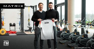 DFB-Athletiktrainer Nicklas Dietrich (rechts) und Volker Lichte (links) stehen im modernen Fitnessraum des DFB-Campus und halten gemeinsam ein weißes Deutschland-Trikot, oben auf der Rückseite der schwarze Aufdruck in Großbuchstaben 'Matrix'. Im Hintergrund sind Trainingsgeräte von Matrix Fitness zu sehen.