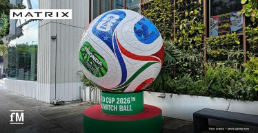 Vor einem Gebäude steht im Vordergrund ein großer Fußball auf einem grünroten Sockel mit den lesbaren Aufschriften „TRIONDA“, „FIFA“ und „WORLD CUP 2026™ OFFICIAL MATCH BALL“; rechts befinden sich Pflanzen, eine Sitzbank und ein Fenster mit Plakaten.