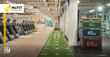 Grüne Kunstrasenbahn mit aufgestapelten Sprungboxen und Laufbändern in einem Fitnessstudio. Links befindet sich das McFIT-Logo.