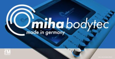 Nahaufnahme eines EMS-Bediengeräts mit Display und mehreren Drehknöpfen; darüber groß eingeblendetes Logo 'miha bodytec – made in germany'; links unten kleines fM-Logo; blau-graue Farbgebung.