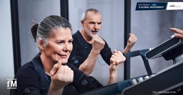 Zwei ältere Erwachsene trainieren mit EMS-Anzügen an einem Trainingsgerät und führen kraftvolle Armbewegungen aus