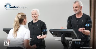Zwei Personen trainieren in EMS-Anzügen an Fitnessgeräten, während eine Trainerin die Übungen betreut