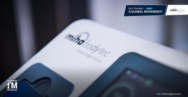 Nahaufnahme eines EMS-Geräts von 'miha bodytec'. Auf der Oberfläche steht 'made in Germany'. Oben rechts ist ein Banner mit dem Slogan 'A Global Movement!' zu sehen.