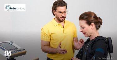 Ein Trainer begleitet eine Frau während eines EMS-Ganzkörpertrainings. Links ist ein EMS-Steuergerät sichtbar.