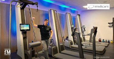 Dirk Rienesl steht in einem modernen Fitnessraum neben einem milon Trainingsgerät mit Bildschirm. Der Raum ist hell, mit Holzverkleidung an der Wand und blauem Licht. Im Hintergrund sind weitere Geräte und Kettlebells zu sehen.