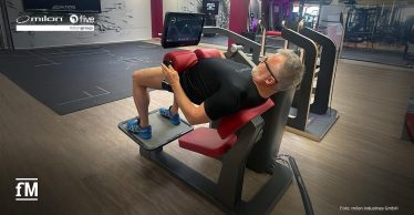 Ein Trainingsgerät vom Typ milon Q+ Hip Thrust in einem Fitnessstudio mit einer trainierenden Person, frontal aufgenommen, gut sichtbar sind Rückenpolster und Display.