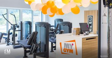 Empfangsbereich eines modernen Fitnessstudios mit Trainingsgeräten, Computerterminal und orange-weißen MYGYM-Luftballons an der Decke.
