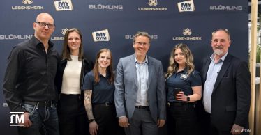 Sechs Personen stehen nebeneinander vor einer dunklen Markenwand mit Logos von MYGYM, LEBENSWERK und BLÜMLING; mittig ein Mann im Anzug, daneben Frauen und Männer in Business- und Polokleidung