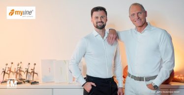 Eric Herrforth (links) und Alexander Dillmann stehen nebeneinander in einem hell ausgeleuchteten Raum mit weißer Wand. Beide tragen weiße, langärmelige Hemden. Dillmann lehnt mit dem rechten Arm auf der Schulter von Herrforth und lächelt in die Kamera. Vor ihnen steht ein Sideboard mit mehreren Trophäen und Ordnern. Links oben im Bild ist das orangefarbene Logo von myline zu sehen, rechts unten der Fotohinweis 'Foto: myline Deutschland GmbH'.