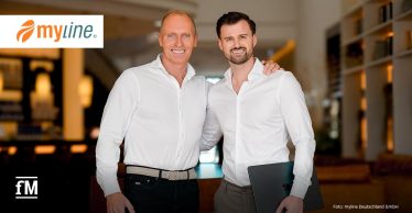 Zwei Männer in weißen Hemden stehen mittig in einem Innenraum; der linke legt den Arm um die Schulter des rechten, der rechte hält einen dunklen Laptop unter dem Arm. Oben links das Logo „myline“, unten links das Logo „fM“, unten rechts der Schriftzug „Foto: myline Deutschland GmbH“; im unscharfen Hintergrund Regale und warmes Licht.