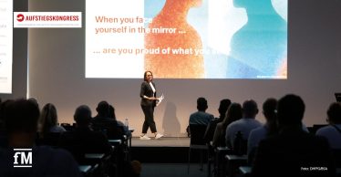 Referentin steht vor großer Leinwand mit Spiegelmotiv und englischem Zitat, Publikum sitzt im abgedunkelten Vortragssaal.