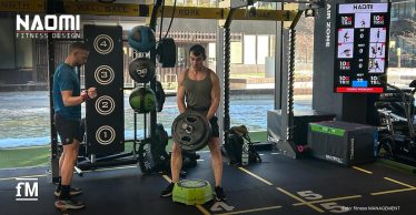 Functional Rig mit Medizinbällen und nummerierter Zielwand, zwei Männer trainieren mit Gewichten auf markierten Bodenfeldern, rechts ein vertikaler 10XTraining-Bildschirm.