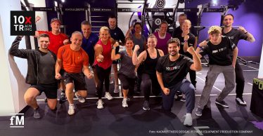 Gruppe von etwa fünfzehn Personen kniet und steht in einem Fitnessstudio und posiert lachend vor einem Trainingsrack.