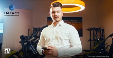 Mann im weißen Hemd hält Smartphone, im Hintergrund Fitnessgeräte und Firmenlogo Impact Unternehmensberatung.