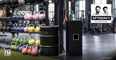 Moderne Fläche im Fitnessstudio in dunklen Farbtönen. Links auf einem Regal liegen bunte kettlebells, rechts steht ein schawrzer Spender für Desinfektionstücher von OPTIMUM11.