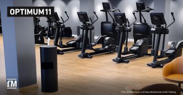 In einem modernen Fitnessstudio stehen Cardiogeräte und ein Spender für Desinfektionstücher. Oben links ist das Logo von Optimum11.