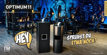 Zwei schwarze Spendersysteme im Vordergrund eines Fitnessstudios mit Kraftgeräten, rechts ein zylindrischer Spender und links ein eckiger Spender, im Hintergrund Trainingsgeräte und Wandgrafik mit muskulöser Figur, auf dem Boden Holzoptik, sichtbare Textelemente „HEY“ und „SPRÜHST DU ETWA NOCH?“.