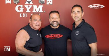 Drei Männer posieren vor einer roten Wand mit Powerhouse-Gym-Logo; einer trägt ein Panatta-T-Shirt.