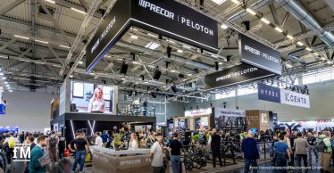 Großer Messestand mit Deckenkonstruktionen und Beleuchtung, mehrfach sichtbare Logos „PRECOR | PELOTON“, viele Besucher zwischen Cardiogeräten und Präsentationsflächen.