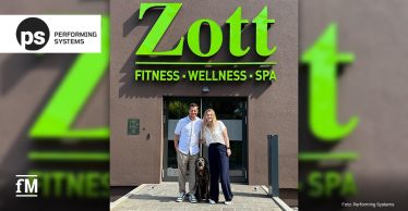 Vor dem Eingang eines Gebäudes mit grünem Schriftzug „Zott FITNESS · WELLNESS · SPA“ stehen ein Mann und eine Frau neben einem schwarzen Hund. Im linken Hintergrund ist das Logo von Performing Systems zu sehen.