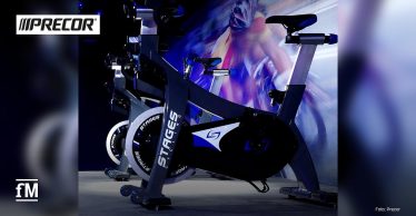 Indoor-Cycling-Bikes von Stages im Ausstellungsraum, beleuchtet mit blauen und weißen Akzenten. Logo von Precor und Stages sind sichtbar.