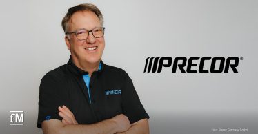 Ein lächelnder Mann mit Brille und kurzen grauen Haaren trägt ein schwarzes Poloshirt mit Precor-Logo. Er steht vor einem neutralen Hintergrund, während das Precor-Logo rechts von ihm zu sehen ist.