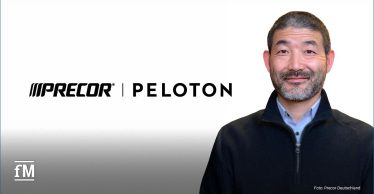 Rechts im Bild eine stehende Person (Precor Präsident Ean Reves,) frontal zur Kamera, neutraler Hintergrund, links daneben großflächig das Precor | Peloton Logo, unten links kleines fM-Logo, unten rechts Bildcredit, Person trägt dunkle Jacke, Oberkörperaufnahme, Blickrichtung nach vorn.