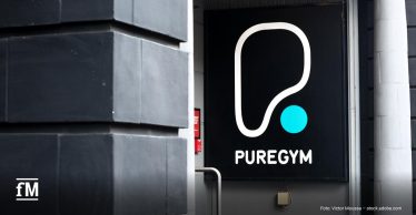 Weißes PureGym-Logo mit türkisfarbenem Punkt auf einer schwarzen Werbetafel am Gebäudeeingang.