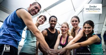 Sechs sportlich gekleidete junge Erwachsene in einem Fitnessstudio stehen im Halbkreis und legen motiviert ihre Hände übereinander, während sie in die Kamera lächeln.