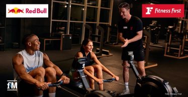 Zwei Personen trainieren auf Rudergeräten in einem warm beleuchteten Fitnessstudio, daneben steht ein Trainer und spricht ihnen motivierend zu. Oben links ist das Red-Bull-Logo, oben rechts das Fitness-First-Logo.