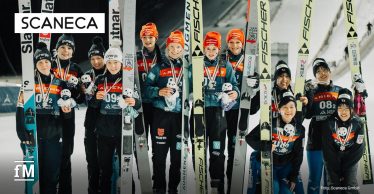 Elf Skispringerinnen posieren in drei nationalen Gruppen für ein Siegerfoto; einige tragen rote Mützen, andere schwarze. Links oben ist das Logo von SCANECA.
