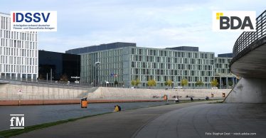 Uferpromenade an einem Fluss mit moderner Büroarchitektur im Hintergrund, links das Logo des „DSSV Arbeitgeberverband deutscher Fitness- und Gesundheits-Anlagen“, rechts das Logo der „BDA“, unten links der Schriftzug „fM“, unten rechts der Bildnachweis „Foto: Stephan Dost – stock.adobe.com“.