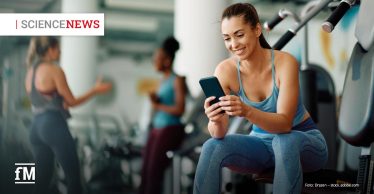 Frau im Sportoutfit sitzt im Fitnessstudio auf einem Trainingsgerät und lächelt beim Blick auf ihr Smartphone.