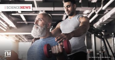 Ein älterer Mann mit grauem Vollbart trainiert in einem Fitnessstudio mit roten Kurzhanteln, während ein jüngerer Trainer in ärmellosem Shirt hinter ihm steht und ihn unterstützt.