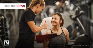 Fitnesstrainerin steht neben sitzendem weiblichem Mitglied mit Hantel, hält Klemmbrett und Stift, beide befinden sich in einem Fitnessstudio.