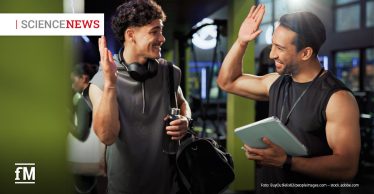 Im Vordergrund begrüßen sich zwei Männer in Sportkleidung mit erhobenen Händen zu einem High Five in einem Fitnessstudio. Die Person links trägt Kopfhörer um den Hals, hält eine Trinkflasche und eine Sporttasche. Die Person rechts hält ein Tablet in den Händen. Im linken oberen Bildbereich steht der Schriftzug „SCIENCE NEWS“, links unten das Logo „fM“.