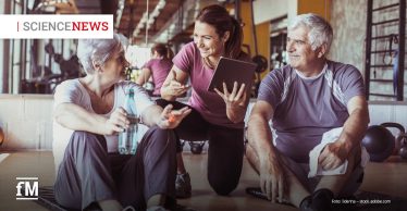 Zwei ältere Menschen in Sportkleidung sitzen in einem Fitnessstudio auf dem Boden und unterhalten sich lächelnd mit einer jüngeren Trainerin, die ein Tablet in der Hand hält.