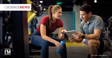 Trainer und Trainierende sitzen im Fitnessstudio nebeneinander und betrachten gemeinsam ein Tablet, Trainingsgeräte im Hintergrund.