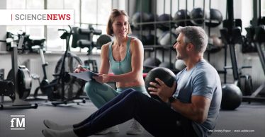 Trainerin sitzt im Fitnessstudio neben einem Mann, der im Sitzen einen Medizinball hält, im Hintergrund Trainingsgeräte.