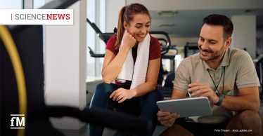 Eine junge Frau mit langem braunen Haar und weißem Handtuch um den Nacken sitzt in Sportkleidung auf einer Bank im Fitnessstudio. Sie lächelt und schaut auf ein Tablet, das ein Mann neben ihr hält. Der Mann trägt ein beiges Poloshirt mit Namensschild und erklärt ihr freundlich etwas auf dem Bildschirm. Beide wirken konzentriert und gut gelaunt. Im Hintergrund sind Fitnessgeräte und große Fenster zu sehen. Oben links steht „SCIENCENEWS“ in weiß-rotem Schriftzug, unten links ist das „fM“-Logo abgebildet.