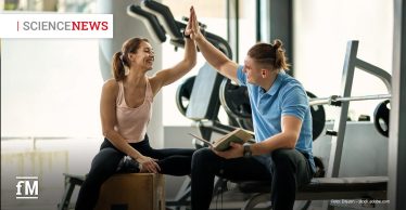 Eine Frau und ein Mann sitzen in einem Fitnessstudio und geben sich lächelnd ein High-Five. Die Frau trägt Sportkleidung und sitzt auf einer Holzkiste, der Mann hält ein Buch in der Hand und sitzt auf einer Hantelbank. Im Hintergrund sind Fitnessgeräte und das Icon 'Science News' zu sehen.