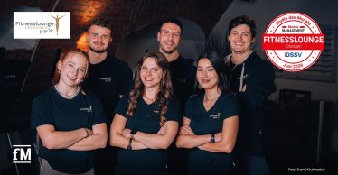 Sechs Mitarbeitende der Fitnesslounge Erlangen stehen in zwei Reihen vor einer Backsteinwand. Alle tragen dunkle Oberteile mit sichtbarem Fitnesslounge-Logo. Oben links ist das Logo der Fitnesslounge sichtbar, rechts das Siegel „Studio des Monats Juni 2026“.