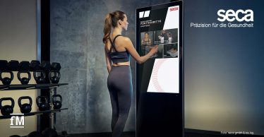 Eine Frau in dunkelgrauem Sportoutfit steht vor einem großen, hochformatigen Touchscreen von seca. Auf dem Bildschirm sind Menüpunkte mit Bildern und Text zu sehen. Links im Bild befinden sich schwarze Kettlebells auf einem Regal vor einer hellen Wand.