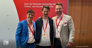 Drei lächelnde Männer mit roten Veranstaltungsbändern stehen nebeneinander vor einer rot-weißen Messewand.