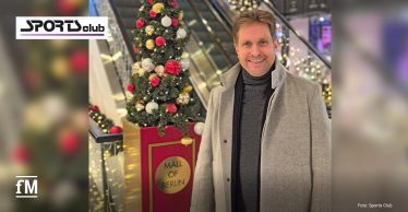 Ein Mann (Sports Club Geschäftsführer Alexander Sosa) in grauem Mantel steht neben dekoriertem Weihnachtsbaum in der Mall of Berlin, im Hintergrund Rolltreppen und Lichterdekoration.