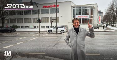 Straßenkreuzung am Saseler Damm in Hamburg-Poppenbüttel mit weißem Eckgebäude und MediaMarkt-Schriftzug, davor steht Alexander Sosa im grauen Mantel auf dem Gehweg.