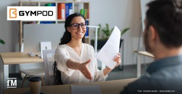 Eine Frau mit Brille sitzt an einem Schreibtisch in einem modernen Büro, reicht einem Mann mit dunklen Haaren, der im Anschnitt von hinten zu sehen ist, die Hand und hält in der anderen Hand Bewerbungsunterlagen. Sie lächelt und zeigt dabei die Zähne. Links oben befindet sich das GYMPOD-Logo auf einer weißen Fläche.