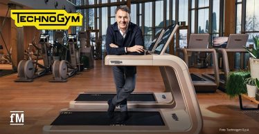 Im Vordergrund steht eine Person (Nerio Alessandri, Technogym Vorsitzender und CEO), die sich mit verschränkten Armen auf ein Laufband stützt, mittig im Raum; im Hintergrund mehrere Fitnessgeräte in Reihen, große Fensterfront und Trainingsfläche sichtbar.