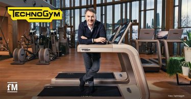 Mann (Technogym CEO Nerio Alessandri) lehnt vorne an einem Technogym-Laufband mit Display, Blick zur Kamera, im Vordergrund Gerät mit Logo „Technogym“, im Hintergrund mehrere Cardiogeräte in einem hellen Fitnessraum mit großen Fenstern, Holzsäulen und Pflanzen.